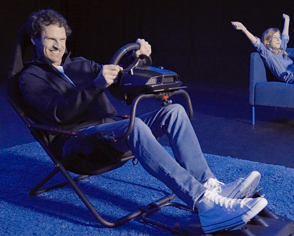 Osoba siedzi w czarnym kokpicie Playseat Challenge Actifit, trzymając kierownicę symulatora. W tle widoczna jest osoba na kanapie z uniesionymi rękami.