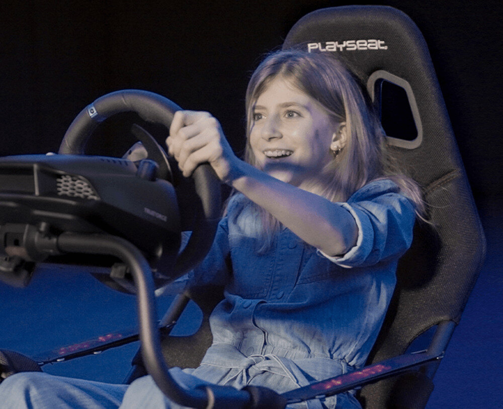 Osoba siedzi w czarnym fotelu Playseat, trzymając kierownicę symulatora jazdy. Na oparciu fotela widoczne jest logo \'Playseat\'.