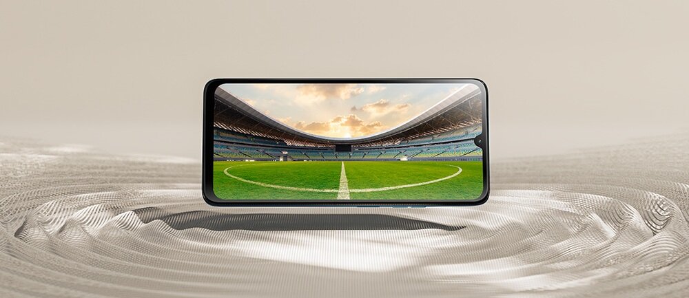 Smartfon wyświetla obraz stadionu piłkarskiego z zieloną murawą i trybunami. Urządzenie jest umieszczone na powierzchni o teksturze przypominającej fale piasku.