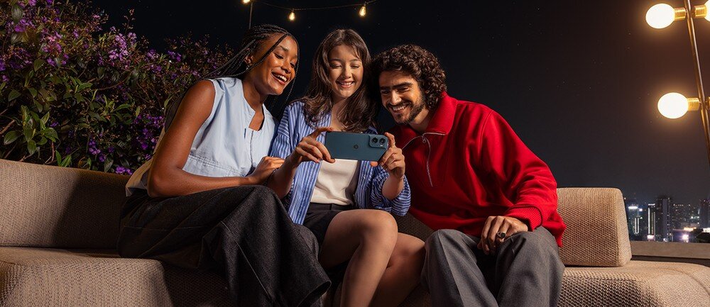 Trzy osoby siedzą na kanapie na zewnątrz, trzymając smartfon i patrząc na jego ekran. W tle widoczne są światła i rośliny.