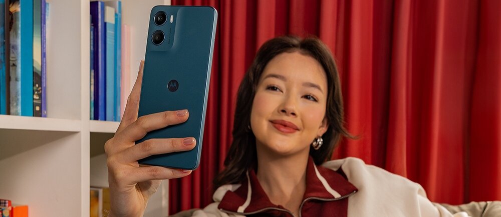 Osoba trzyma niebieski smartfon z logo Motorola, na tle czerwonych zasłon i półki z książkami.