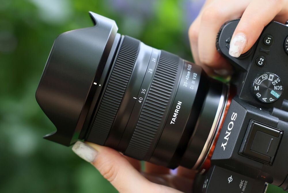 Osoba trzyma aparat fotograficzny z zamontowanym obiektywem TAMRON 20-40mm f/2.8 Di III VXD. Na obiektywie widoczne są oznaczenia ogniskowej i przysłony, a na aparacie logo Sony.