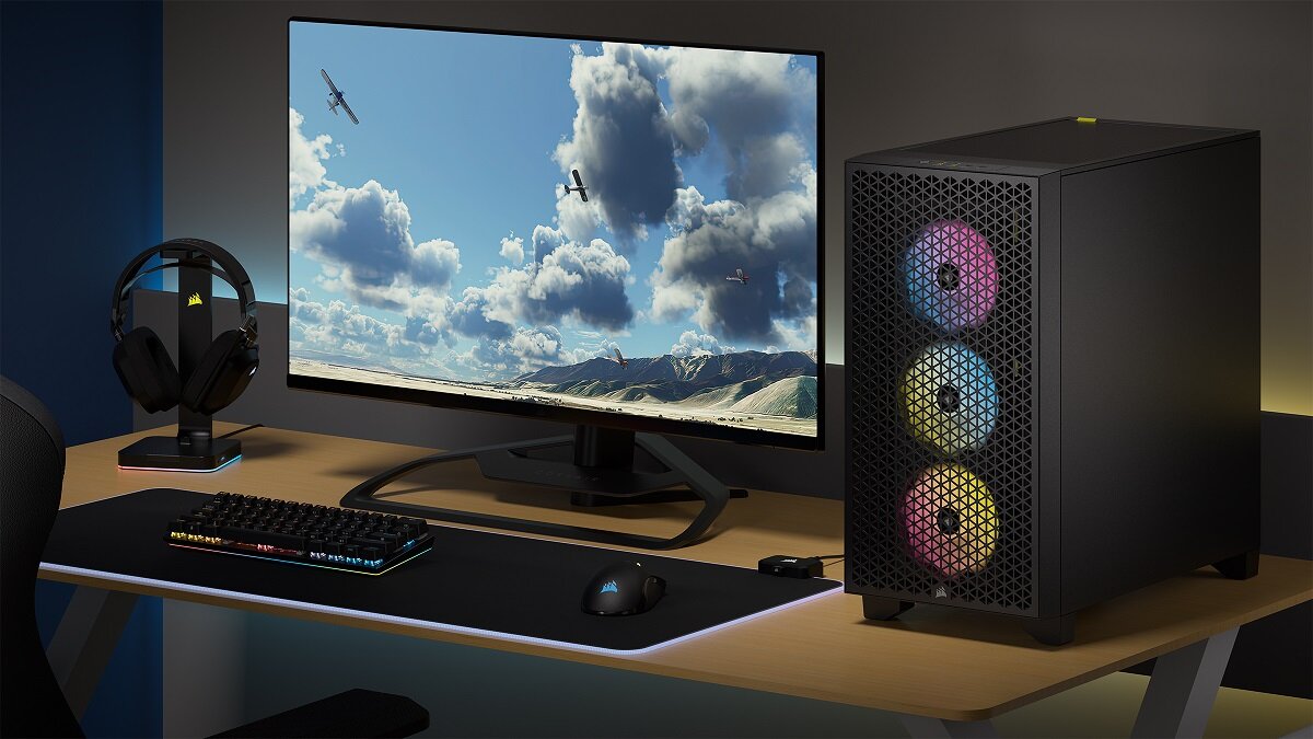 Na biurku znajduje się monitor z wyświetlanym krajobrazem, klawiatura, myszka oraz słuchawki na stojaku. Obok monitora stoi czarna obudowa komputera z trzema podświetlanymi wentylatorami.