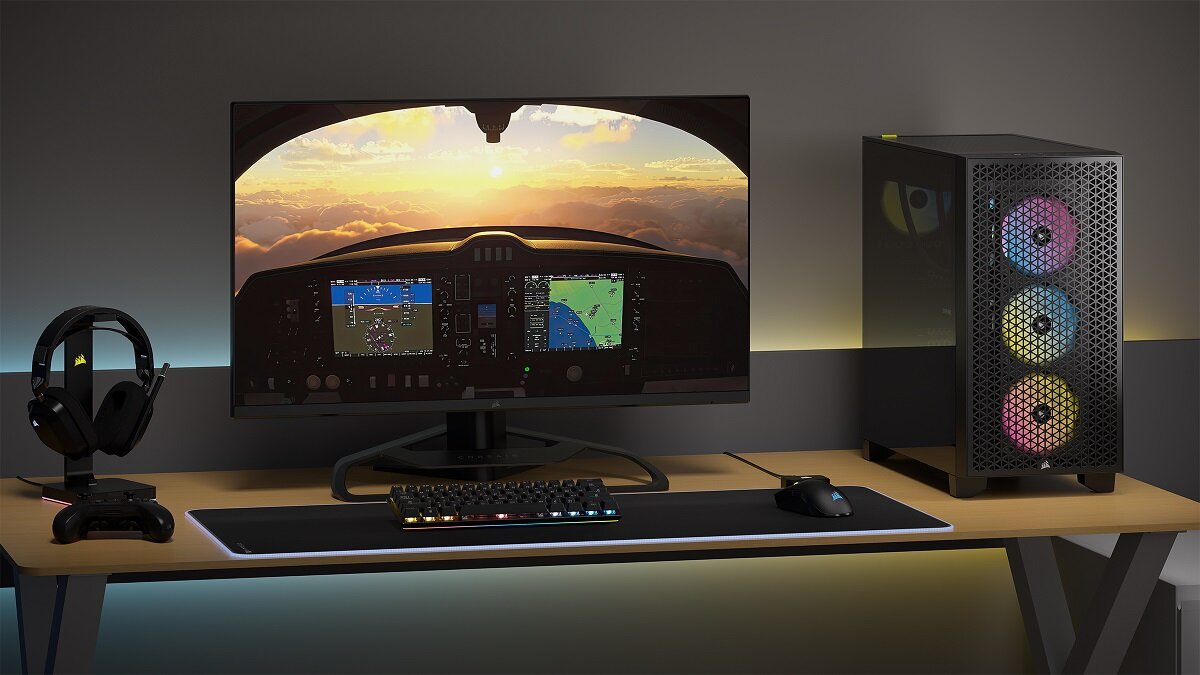 Na biurku znajduje się monitor z symulatorem lotu, klawiatura, myszka, słuchawki oraz obudowa komputera z trzema podświetlanymi wentylatorami. W tle widoczny jest zachód słońca nad chmurami.