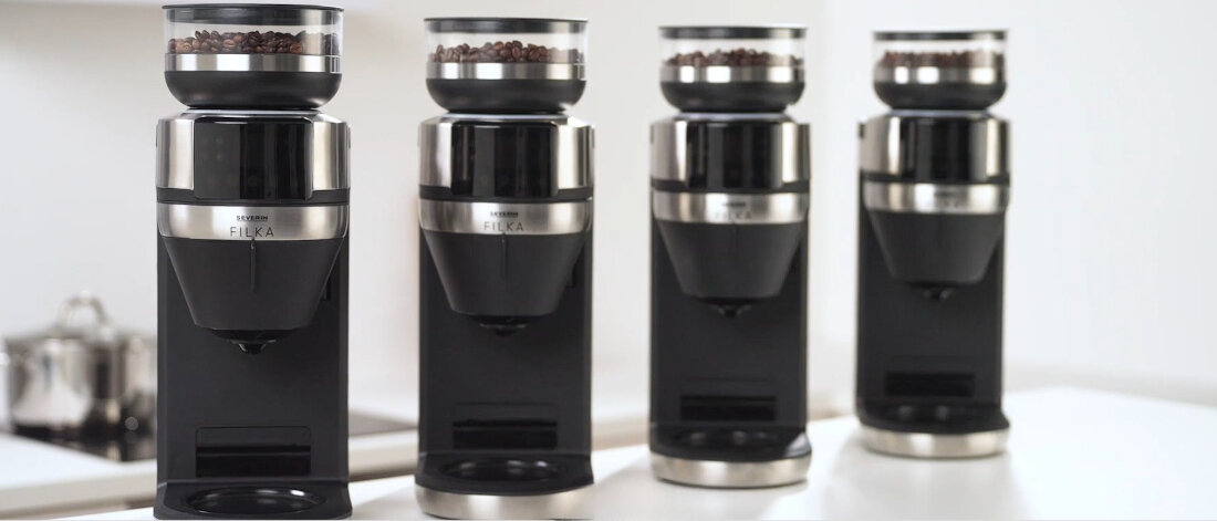 Na obrazie widoczne są cztery ekspresy do kawy z napisem \'FILKA\' na metalowej części, każdy z pojemnikiem na ziarna kawy na górze. Ekspresy stoją na białej powierzchni w jasnym pomieszczeniu.
