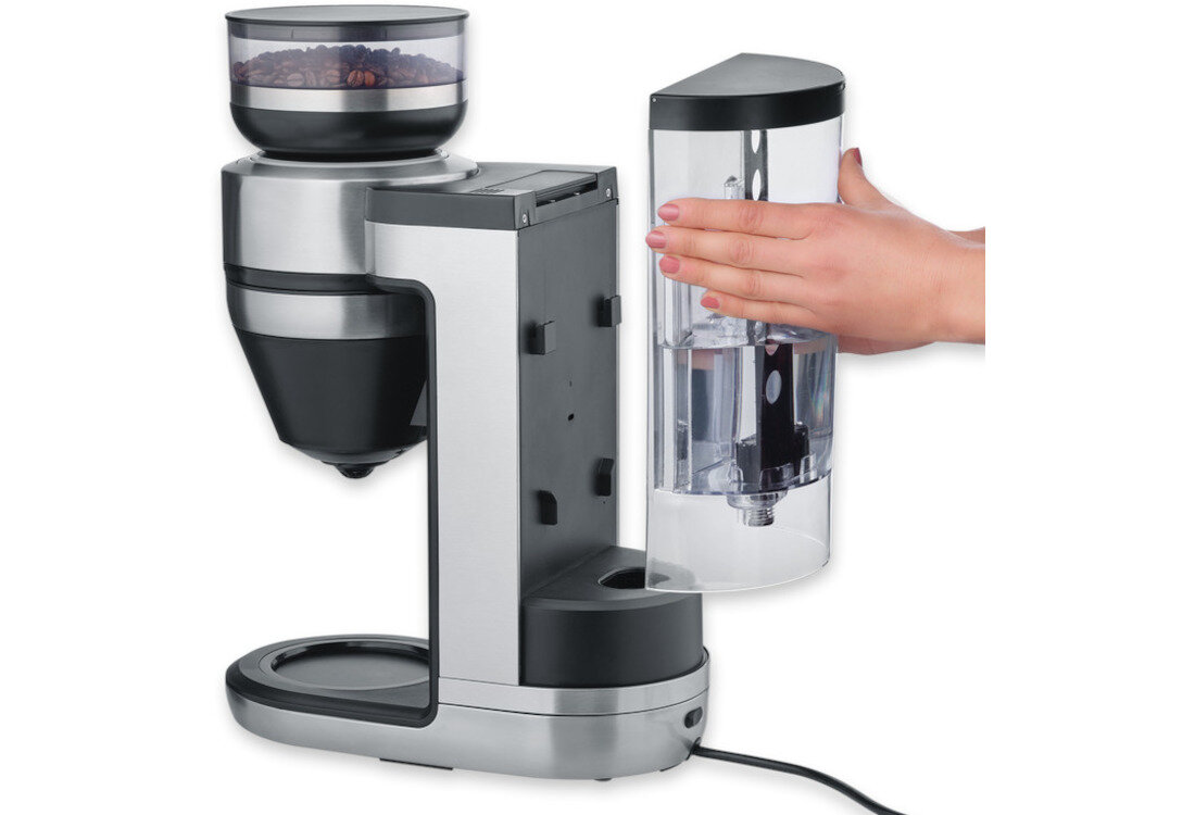 Na obrazie widoczny jest ekspres do kawy z przezroczystym pojemnikiem na wodę, który jest trzymany przez rękę. Ekspres ma metaliczne wykończenie i wbudowany młynek z ziarnami kawy.