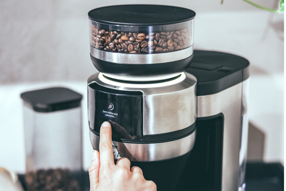 Ekspres do kawy z przezroczystym pojemnikiem na ziarna kawy, na którym widoczny jest tekst: \'Dein Kaffee wird zubereitet...\'.