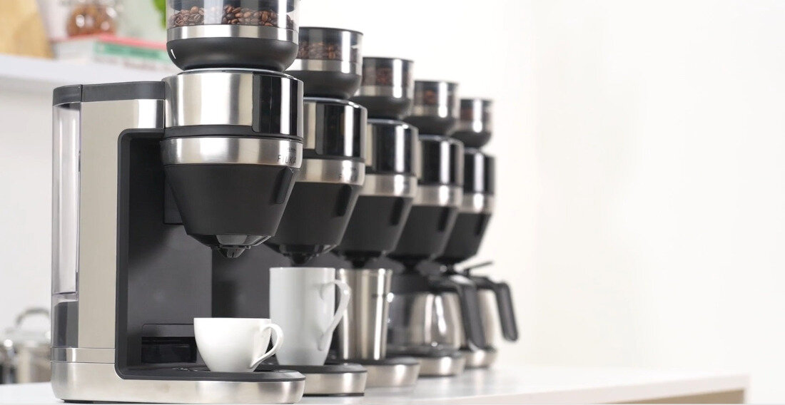 Na obrazie widoczne są ekspresy do kawy ustawione w rzędzie, z filiżankami umieszczonymi pod wylewkami. Ekspresy mają metaliczne wykończenie i przezroczyste pojemniki na ziarna kawy.