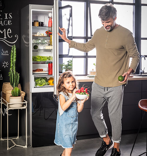 Otwarta lodówka z widocznymi półkami pełnymi jedzenia, obok stoi mężczyzna trzymający owoc i dziecko z miską truskawek. W tle widać kuchnię z dużym oknem i tablicą z napisem \'This is my happy place\'.