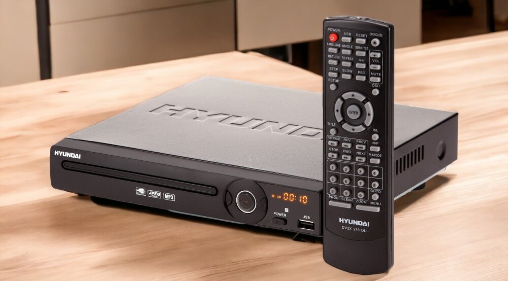 Na drewnianym stole znajduje się odtwarzacz DVD marki Hyundai z wyświetlaczem pokazującym \'00:10\'. Obok leży pilot z wieloma przyciskami, również oznaczony logo Hyundai i napisem \'DV2-X 279 DU\'.