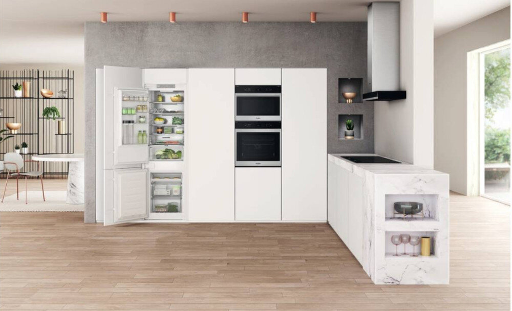 Nowoczesna kuchnia z białymi szafkami, w której znajduje się otwarta lodówka z widocznymi produktami spożywczymi. Obok lodówki znajdują się dwa wbudowane piekarniki, a po prawej stronie marmurowy blat z płytą grzewczą i okapem.