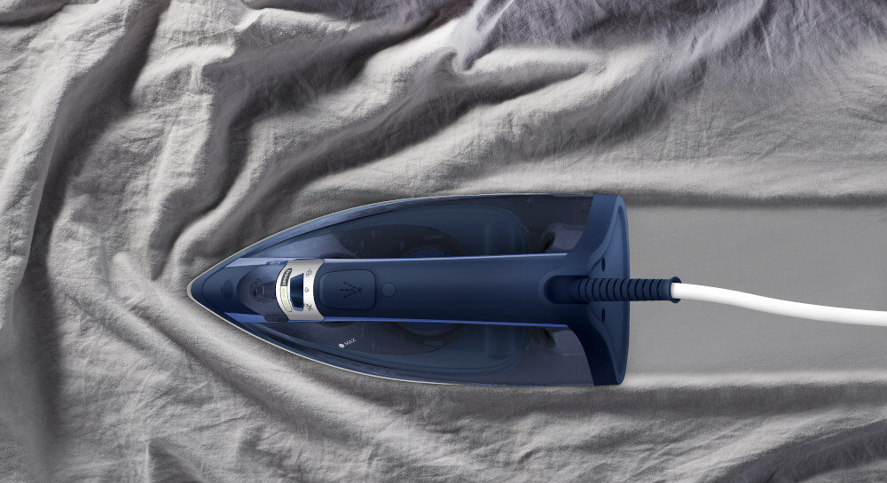 Żelazko w kolorze granatowym przesuwa się po szarej, pogniecionej tkaninie, pozostawiając za sobą wygładzoną powierzchnię. Widoczny jest biały przewód zasilający.