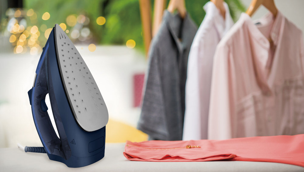 Na pierwszym planie znajduje się żelazko w kolorze granatowym z metalową stopą. W tle widoczne są wyprasowane koszule na wieszakach oraz rozprasowana różowa koszula na desce do prasowania.