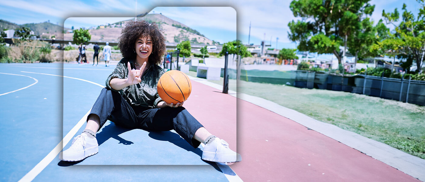 Osoba siedzi na boisku do koszykówki, trzymając pomarańczową piłkę do koszykówki. W tle widać drzewa i wzgórze.