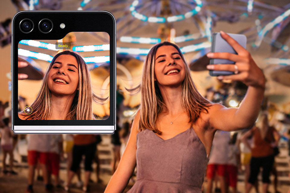 Osoba robiąca selfie smartfonem, na ekranie którego widać jej uśmiechniętą twarz. W tle rozmyte światła i ludzie, a na ekranie telefonu widoczny jest napis \'Ready\'.