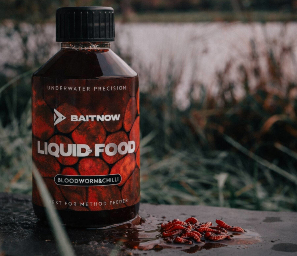 Liquid BAITNOW Food Bloodworm & Chili (250 ml) butelka z liquidem w naturalnym otoczeniu w tle woda obok larwy zalane syropem