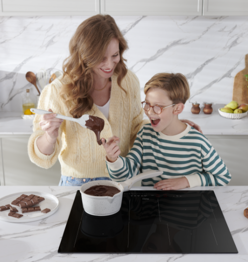 Kobieta trzyma łyżkę z czekoladą nad garnkiem na płycie indukcyjnej. Obok leży tabliczka czekolady.