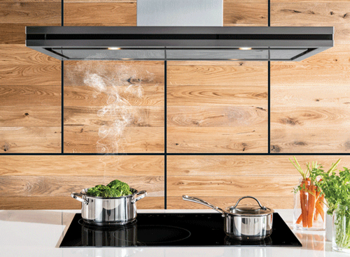 Na zdjęciu widać czarny okap kuchenny zamontowany nad płytą grzewczą z dwoma garnkami. W tle znajduje się drewniana ściana, a obok garnków stoją świeże warzywa.