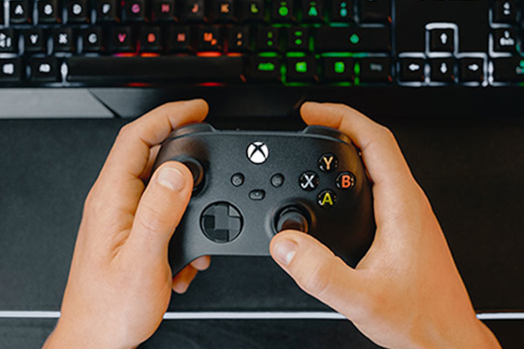 Osoba trzyma czarny kontroler do gier z widocznym logo Xbox. W tle znajduje się klawiatura z podświetleniem RGB.