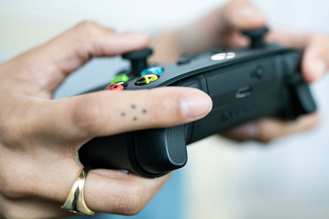 Osoba trzyma czarny kontroler do gier z kolorowymi przyciskami i widocznym logo Xbox. Na palcu widoczne są małe tatuaże w formie kropek.