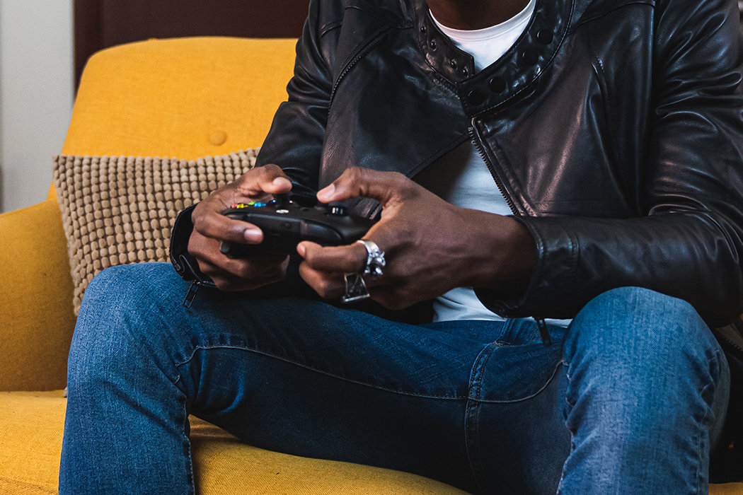 Osoba trzyma czarny kontroler do gier, siedząc na żółtej kanapie. Ubrana jest w skórzaną kurtkę i dżinsy.