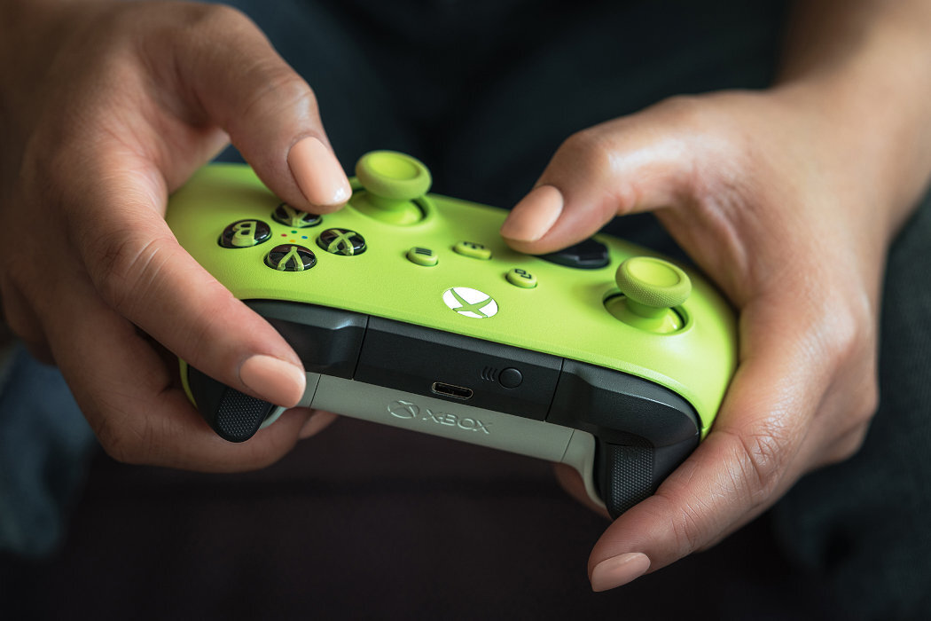 Osoba trzyma zielony kontroler do gier z widocznym logo Xbox. Kontroler ma czarne przyciski i port USB na spodzie.