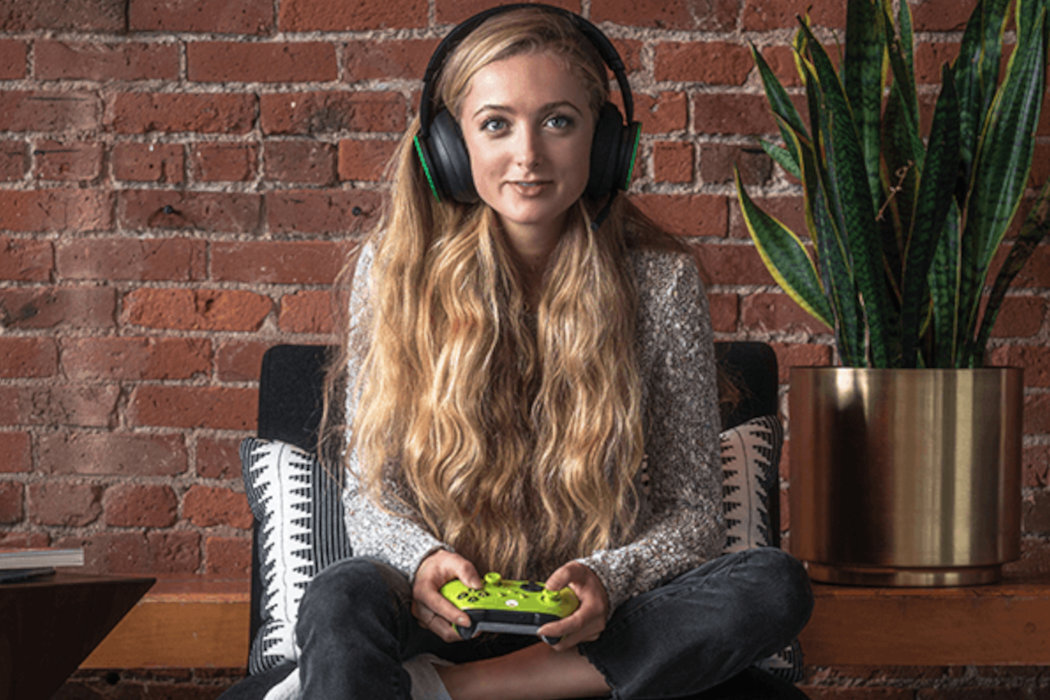Osoba siedzi na kanapie z kontrolerem do gier w kolorze zielonym w rękach, na głowie ma duże słuchawki. W tle widoczna jest czerwona ceglana ściana i roślina w doniczce.