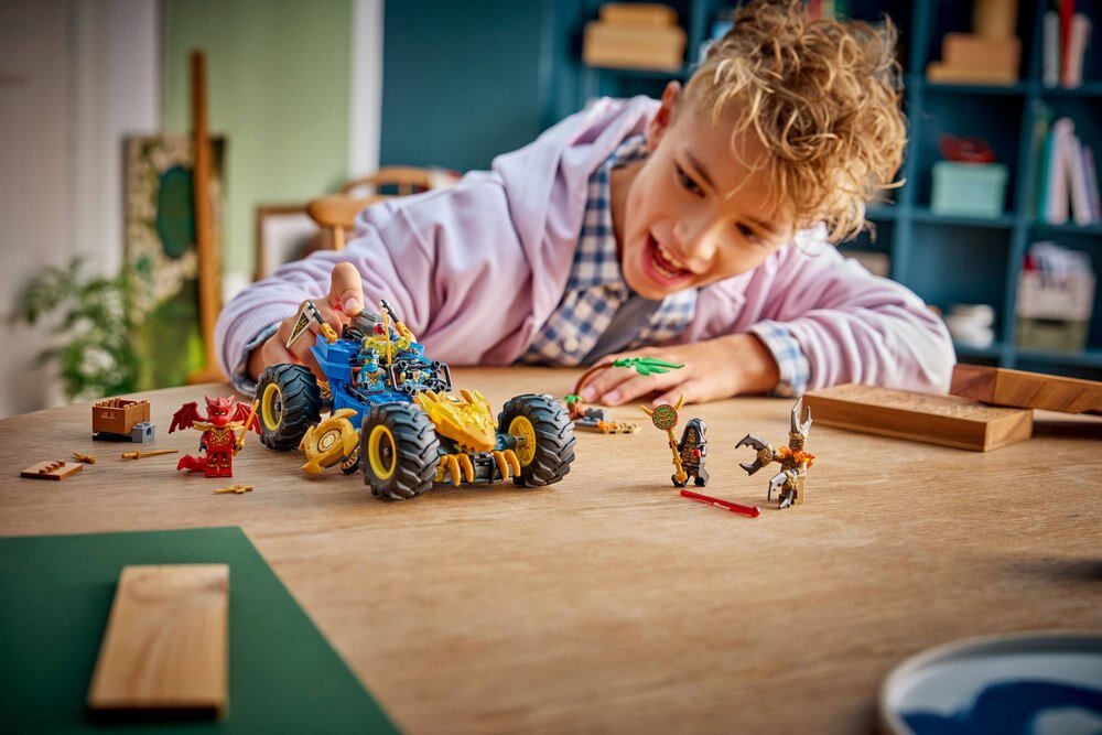 Dziecko bawi się zestawem LEGO, w tym dużym, niebiesko-żółtym pojazdem z dużymi kołami oraz kilkoma figurkami. W tle widać półki z książkami i dekoracjami.