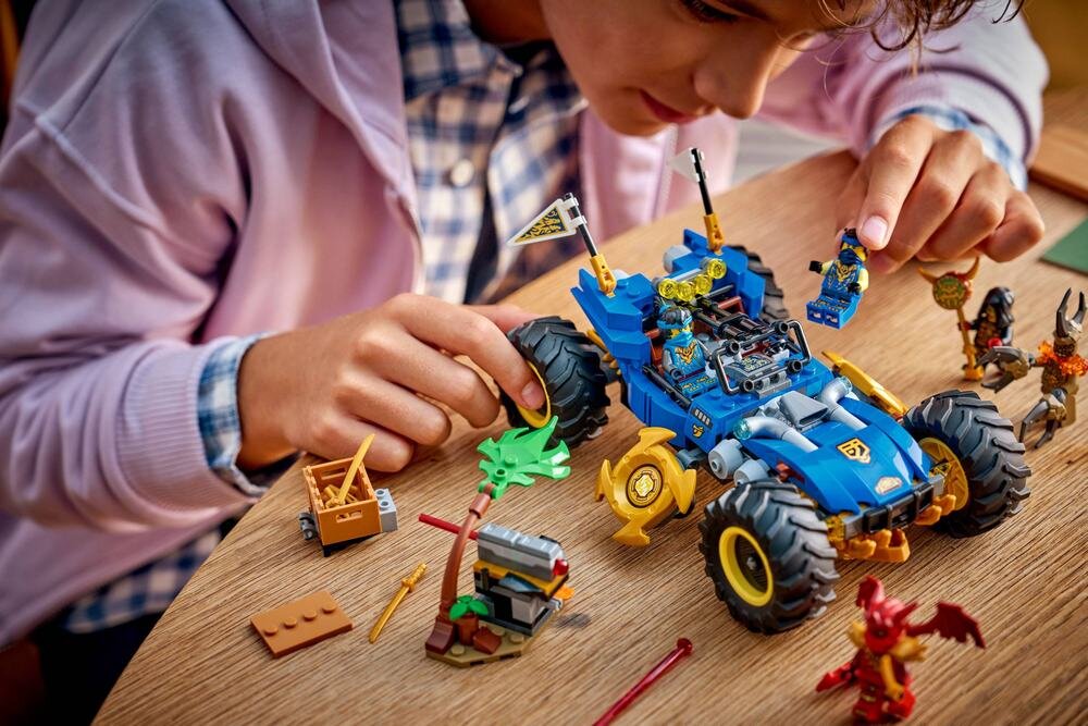 Dziecko bawi się zestawem LEGO, w tym niebieskim samochodem terenowym z dużymi kołami i figurkami. Na stole znajdują się różne elementy konstrukcyjne i figurki LEGO.