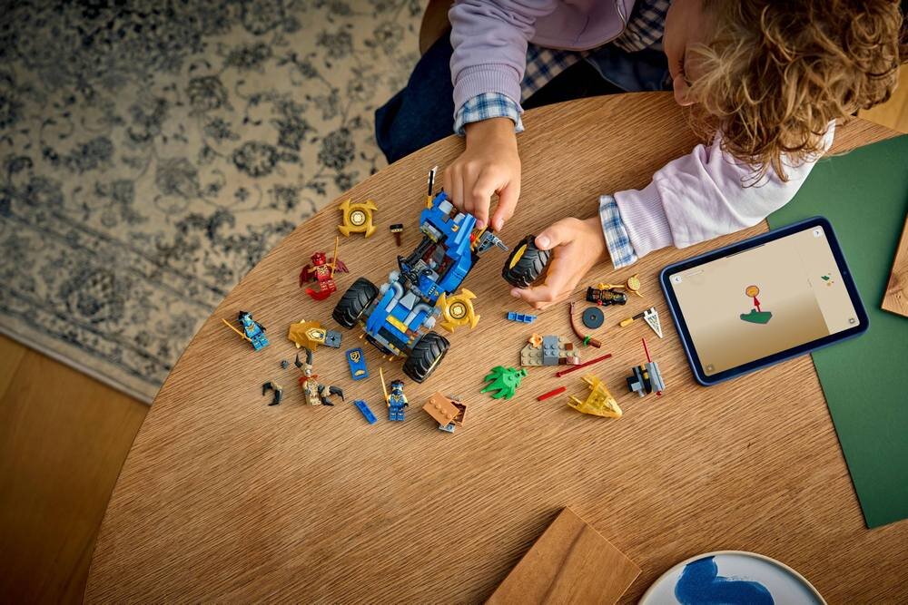 Na drewnianym stole znajduje się zestaw klocków LEGO, w tym niebieski pojazd i kilka figurek. Obok leży tablet z wyświetlonym rysunkiem.