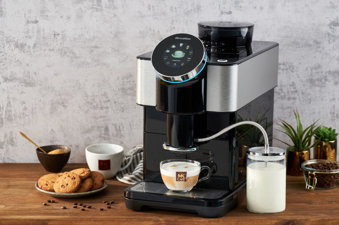 Na drewnianym blacie stoi czarny ekspres do kawy z logo \'Dr.Coffee\', obok filiżanka z napisem \'MK Cafe\' oraz szklany pojemnik z mlekiem. W tle widoczne są filiżanki, talerz z ciastkami i rośliny doniczkowe.