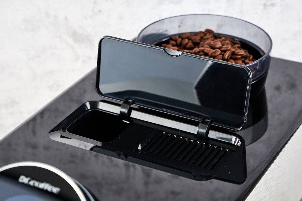 Na obrazie widoczny jest czarny ekspres do kawy z otwartym pojemnikiem na ziarna kawy. Na urządzeniu znajduje się logo \'Dr.Coffee\'.
