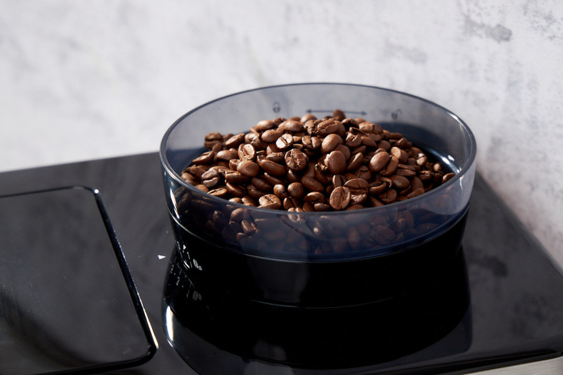 Na obrazie widoczna jest przezroczysta miska wypełniona ziarnami kawy, umieszczona na czarnej powierzchni. W tle znajduje się jasna, teksturowana ściana.