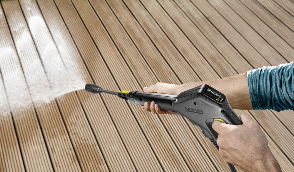 Osoba używa myjki ciśnieniowej do czyszczenia drewnianej powierzchni. Na urządzeniu widoczne jest logo \'KARCHER\'.