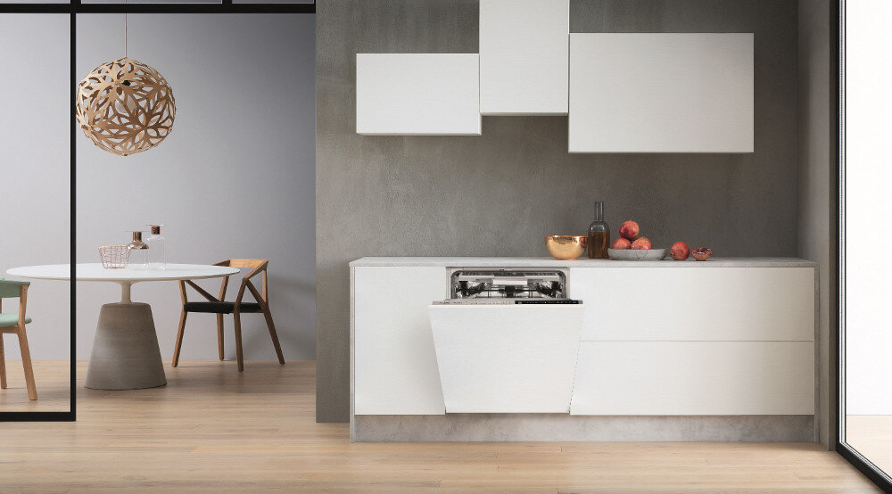 Nowoczesna kuchnia z otwartą zmywarką w zabudowie, obok której znajdują się białe szafki i szare ściany. W tle widoczny jest stół z krzesłami oraz dekoracyjna lampa sufitowa.