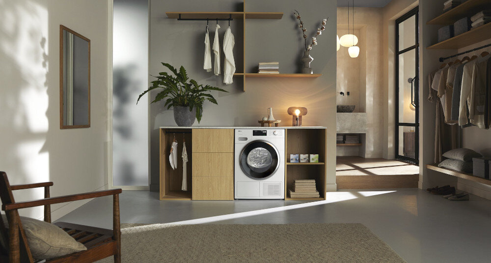 Wnętrze z suszarką umieszczoną w zabudowie meblowej, obok której znajdują się półki z ubraniami i rośliną doniczkową. Na ścianie wisi lustro, a pomieszczenie jest oświetlone lampami sufitowymi.