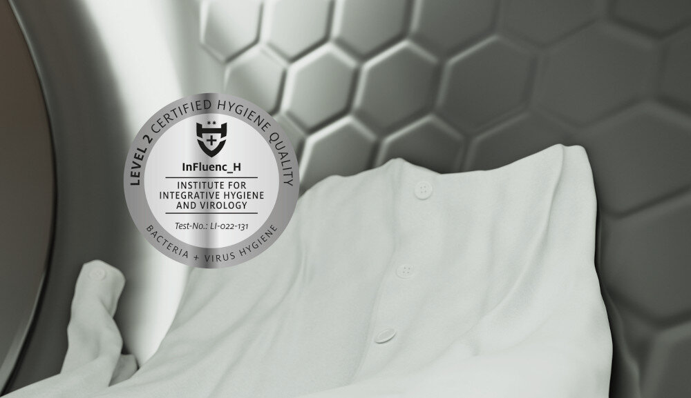 Wnętrze bębna suszarki z widoczną białą tkaniną. Na bębnie znajduje się naklejka z tekstem: \'LEVEL 2 CERTIFIED HYGIENE QUALITY, InFluenc_H, INSTITUTE FOR INTEGRATIVE HYGIENE AND VIROLOGY, Test-No.: LI-022-131, BACTERIA + VIRUS HYGIENE\'.