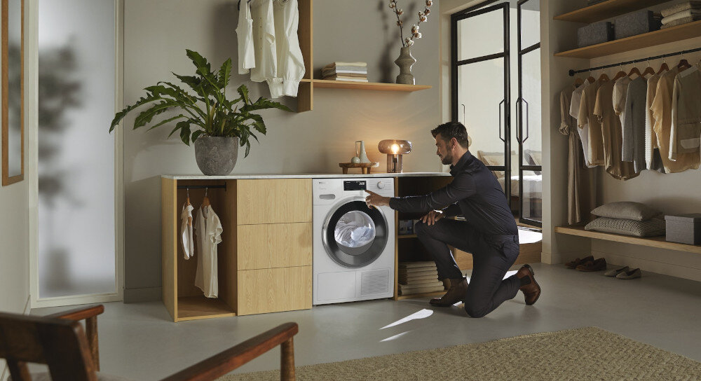 W pomieszczeniu znajduje się suszarka z otwartymi drzwiami, obok której stoi mężczyzna. Na blacie obok suszarki stoi roślina w doniczce.