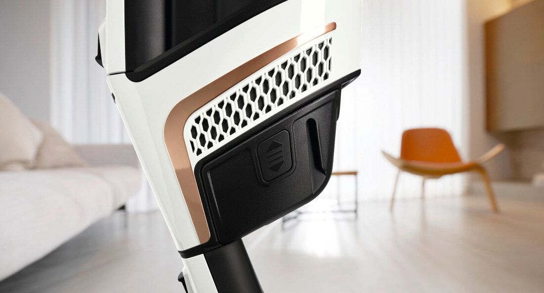 Zbliżenie na część odkurzacza z białą obudową i czarnym elementem z logo. W tle widoczny jest nowoczesny salon z pomarańczowym krzesłem.