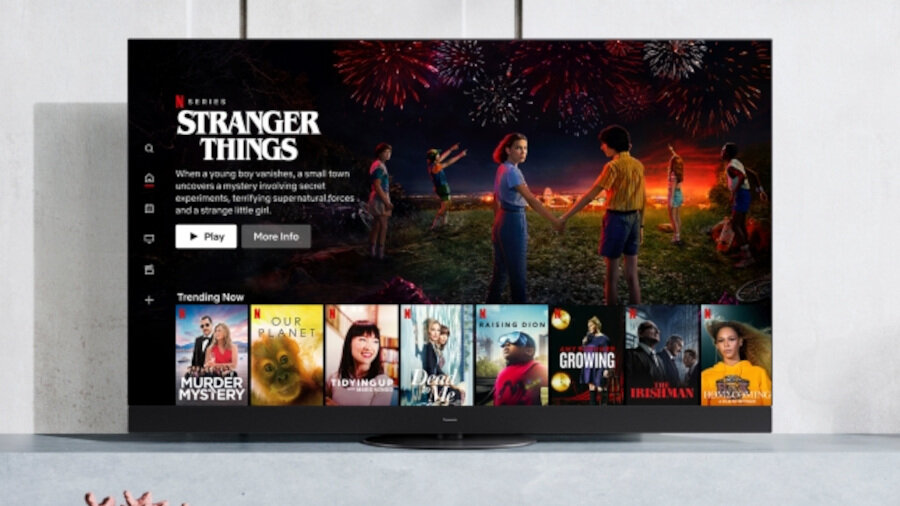 Telewizor wyświetla interfejs Netflix z widocznym tytułem \'Stranger Things\' oraz miniaturami innych filmów i seriali. Na ekranie widać scenę z fajerwerkami i grupą osób na tle zachodzącego słońca.
