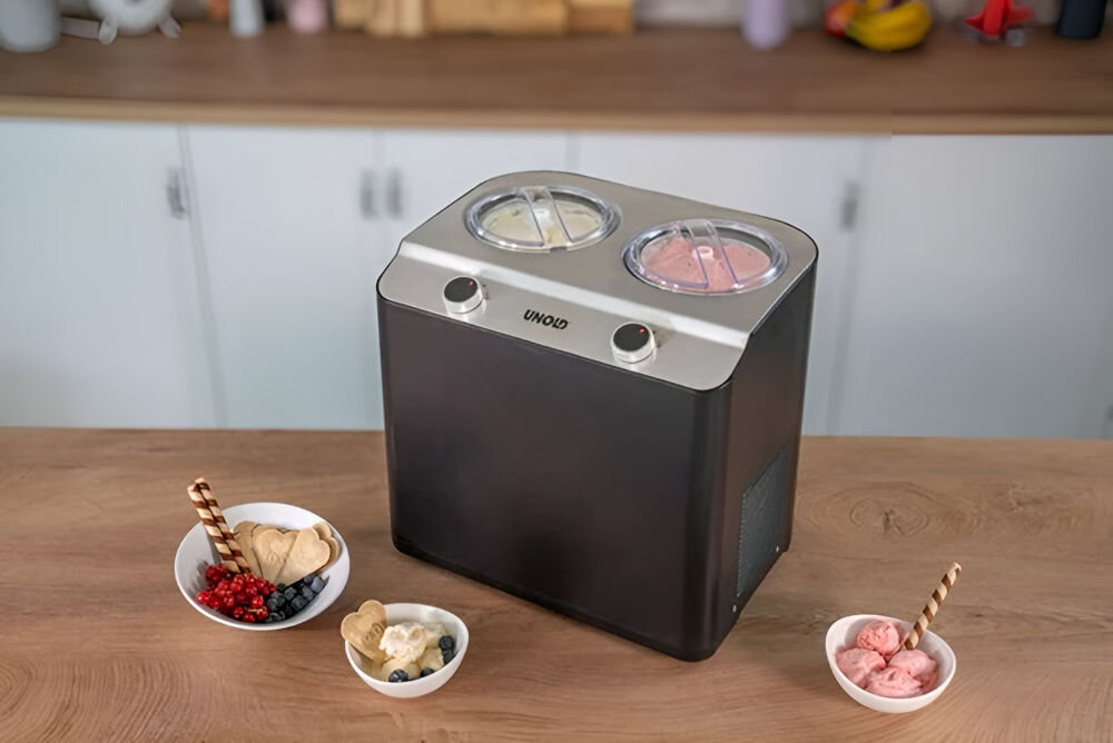 Na drewnianym blacie stoi czarna maszynka do lodów z dwoma przezroczystymi pokrywami na górze, z logo \'UNOLD\'. Obok znajdują się trzy miseczki z lodami i owocami.