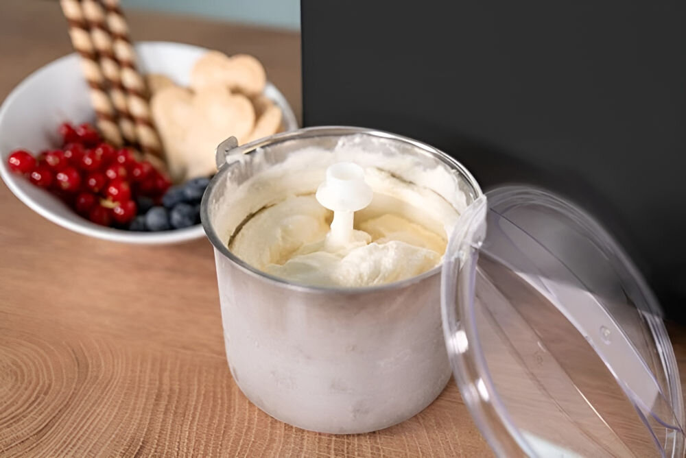 Na drewnianym blacie stoi metalowy pojemnik z białymi lodami, obok znajduje się przezroczysta pokrywka. W tle widoczna jest miska z owocami i wafelkami.