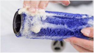Osoba myje niebieski wałek z pianą, trzymając go w dłoniach. W tle widoczny jest rozmyty element odkurzacza.