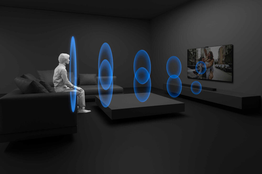 W ciemnym pokoju znajduje się osoba siedząca na kanapie, przed którą na stoliku widoczne są niebieskie fale dźwiękowe. Na ścianie wisi telewizor, a pod nim umieszczony jest soundbar.