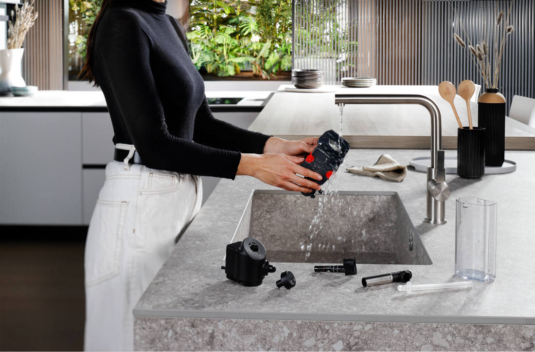 Osoba myje część ekspresu do kawy pod bieżącą wodą w nowoczesnej kuchni. Na blacie obok zlewu znajdują się różne elementy ekspresu.
