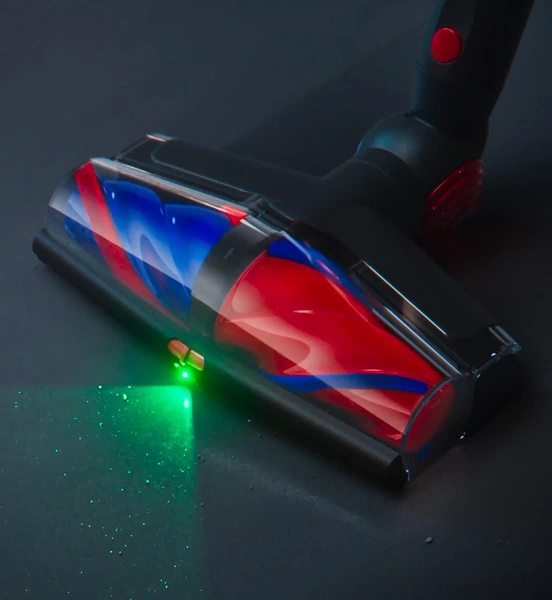 Głowica odkurzacza z przezroczystą obudową, wewnątrz której widoczne są czerwone i niebieskie elementy. Z przodu emitowane jest zielone światło, oświetlające powierzchnię.