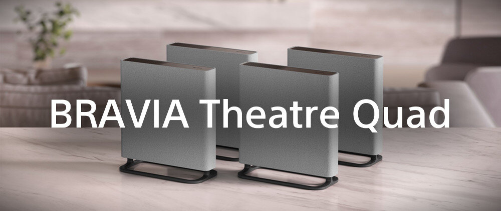 Na obrazie widoczne są cztery głośniki w kolorze czarnym, ustawione na jasnej powierzchni. Na pierwszym planie znajduje się tekst: \'BRAVIA Theatre Quad\'.