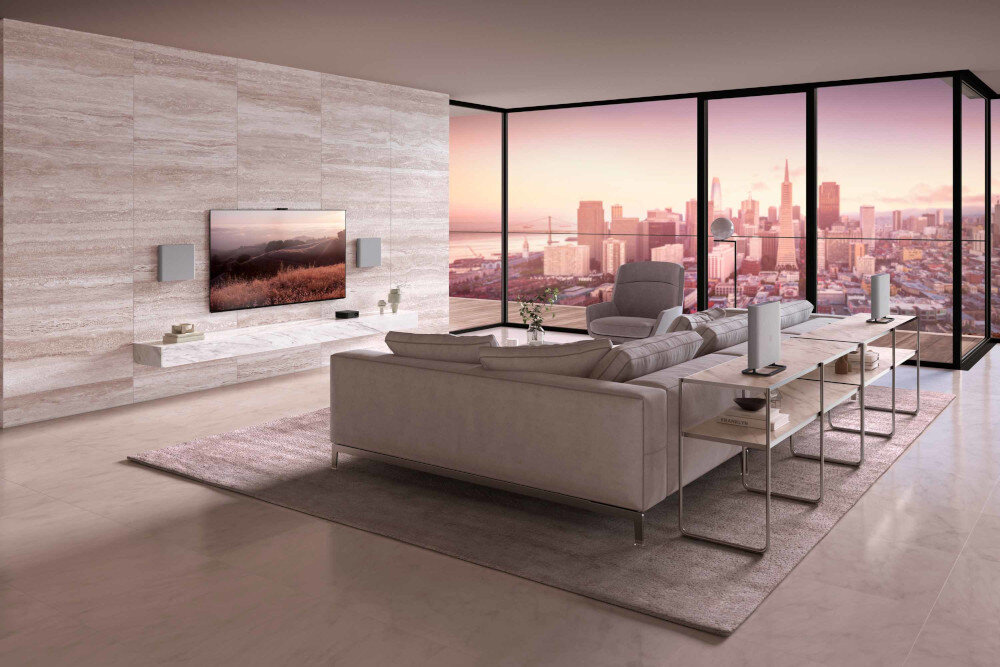 Nowoczesny salon z dużymi oknami, przez które widać panoramę miasta o zachodzie słońca. Na ścianie zamontowany jest telewizor z dwoma głośnikami po bokach, a przed nim stoi jasna sofa i stoliki z dekoracjami.