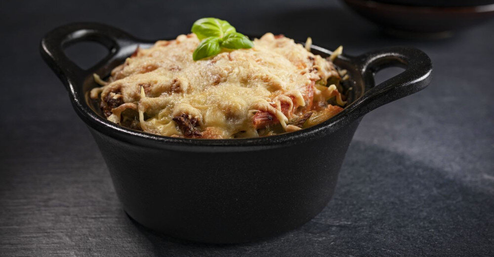 Czarna ceramiczna miska z uchwytami zawiera zapiekankę z serem i liściem bazylii na wierzchu. Miska stoi na ciemnym, kamiennym blacie.