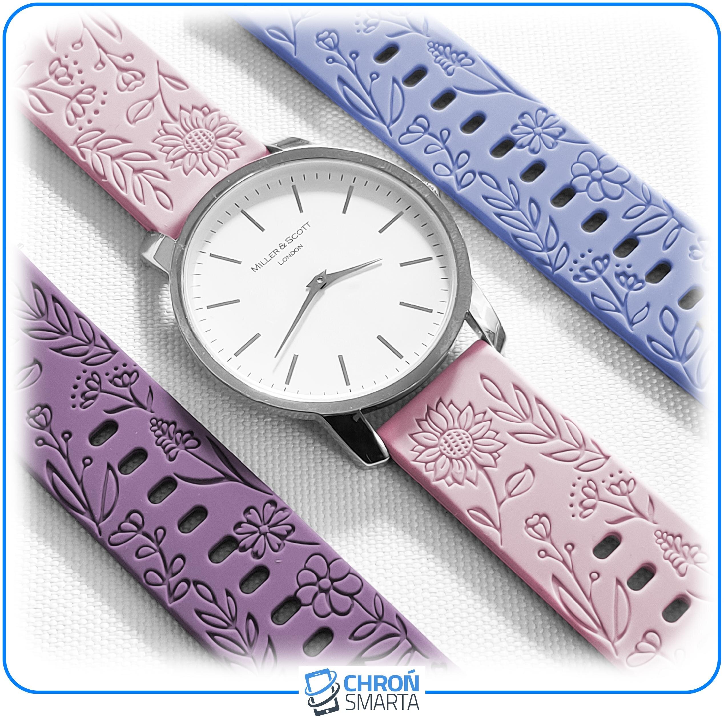 Na obrazie widoczny jest zegarek z białą tarczą i trzema wymiennymi paskami w kolorach różowym, fioletowym i niebieskim, ozdobionymi wzorami kwiatowymi. Na tarczy zegarka znajduje się napis \'Miller & Scott London\'.